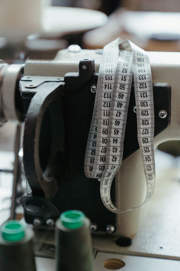 about-us-01 Close-up of a sewing machine with measuring tape in a workshop, highlighting tailoring tools.